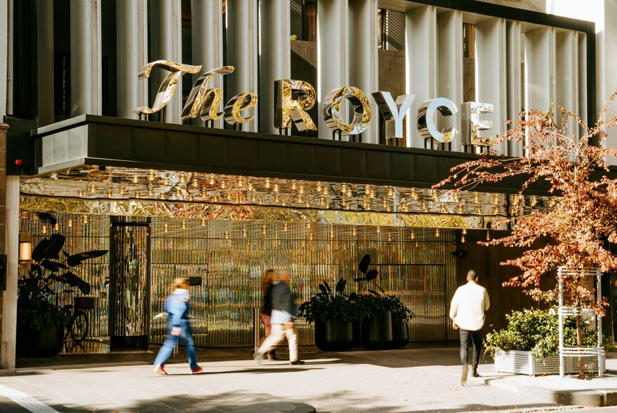 Exterior of a hotel, with people walking past the glass entrance, and signage that reads The ROYCE on the building.