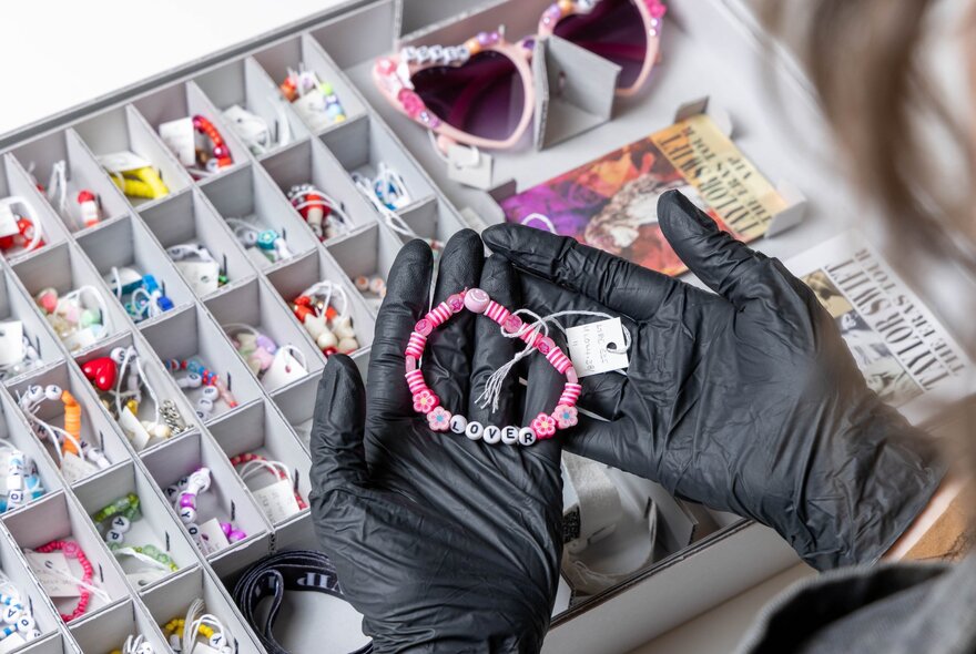 Gloved hands holding beaded bracelet over an archive container of beads.