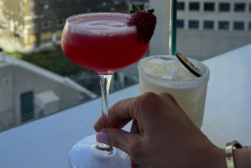A hand holding a red cocktail on a rooftop bar with city views.