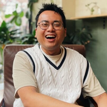 Joshua Hortinela, a smiling Asian man wearing glasses, a beige t-shirt, and a white cable-knit sweater vest with a dark V-neck trim.