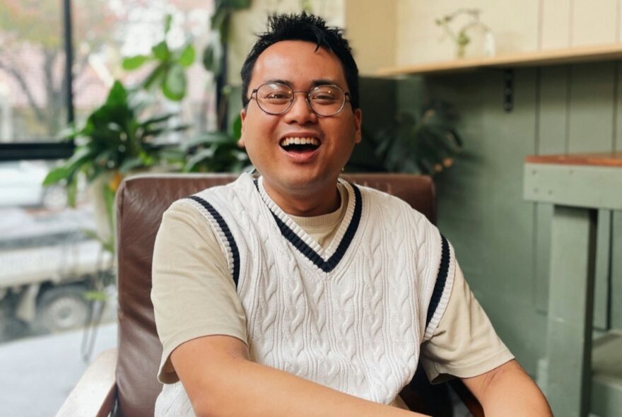 Joshua Hortinela, a smiling Asian man wearing glasses, a beige t-shirt, and a white cable-knit sweater vest with a dark V-neck trim.