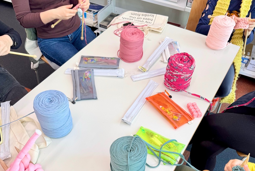 A group of people participating in a knitting craft workshop with spools of brightly coloured yarn, needles, and tools laid out on a table.