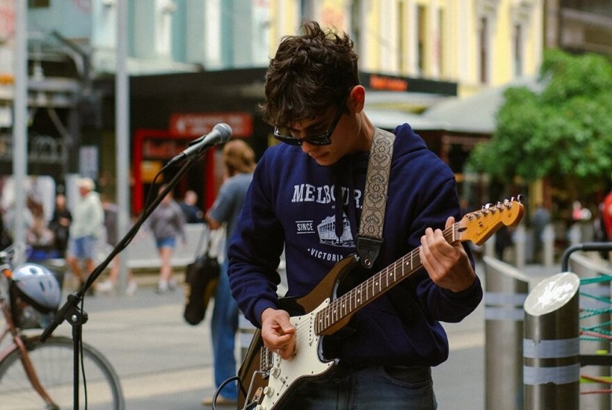 A busker performing in the street playing an electric guitar with his head facing downwards looking at the instrument. 