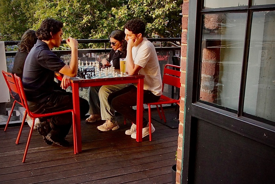 People playing chess at an outdoor table on a wooden deck, with a brick wall and door nearby.