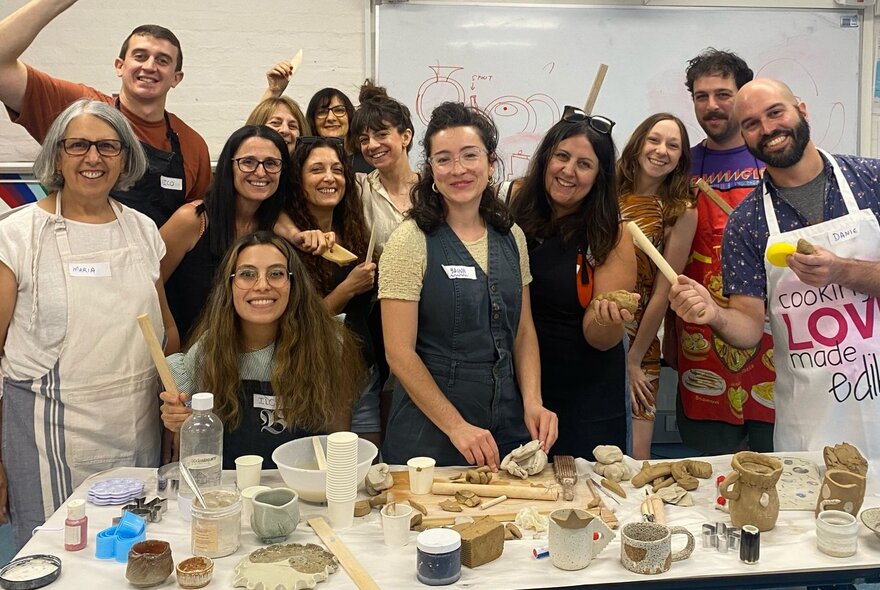 A group of clay workshop participants in a room around a table of clay objects, smiling.