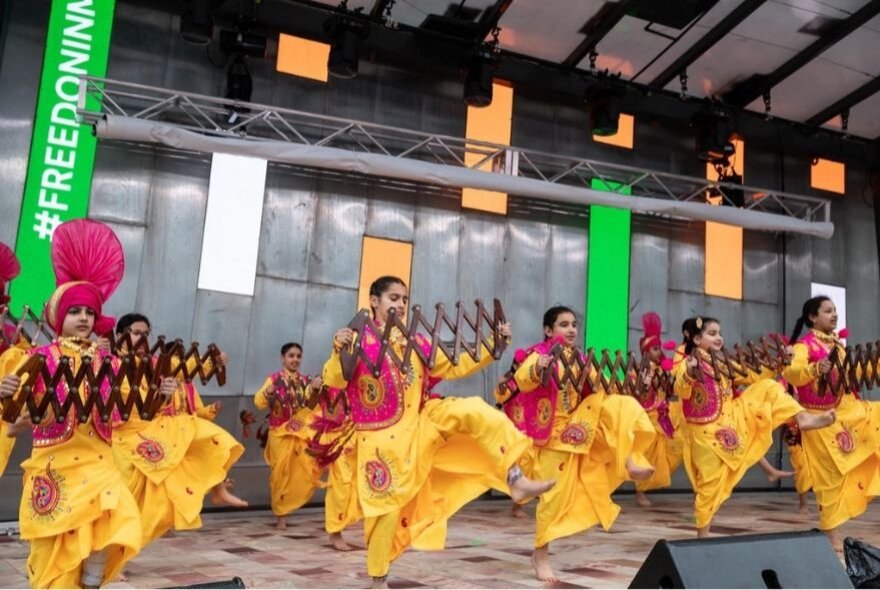 A group of Indian dancers in yellow pants and red tops dancing on a stage in unison. 