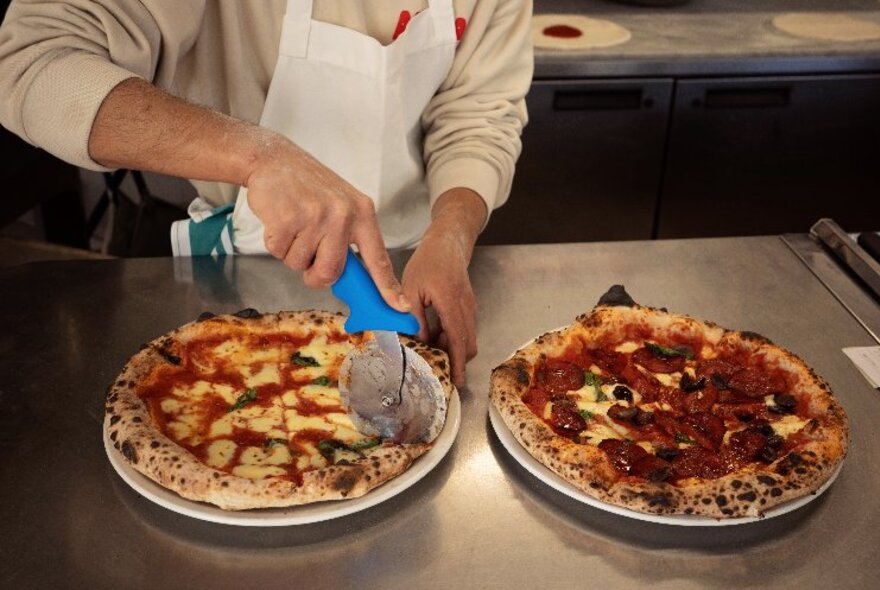 Chef slicing pizzas with a round cutter on a steel bench.