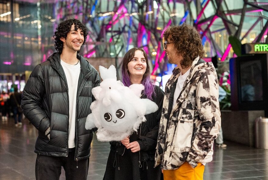 Three people, one with a fluffy white character on a stick, at Fed Square.