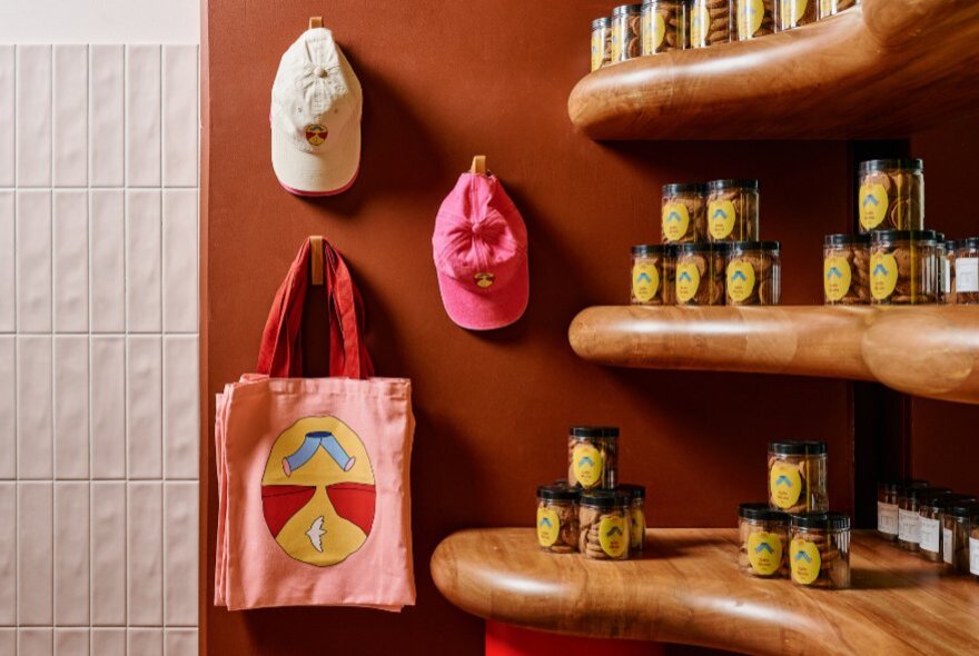 Wooden shelves with jars next to pink tote bags hanging on a hook underneath two hanging hats.