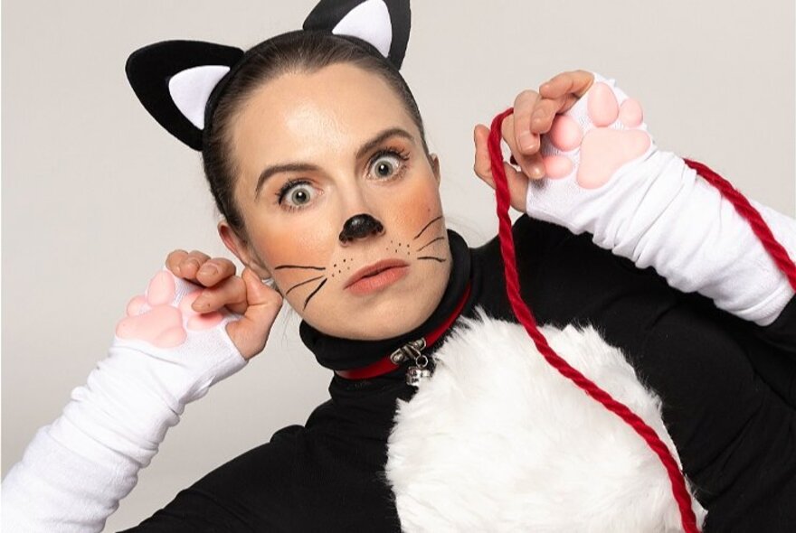 A woman dressed as a black and white cat with a startled expression and holding a piece of red wool.