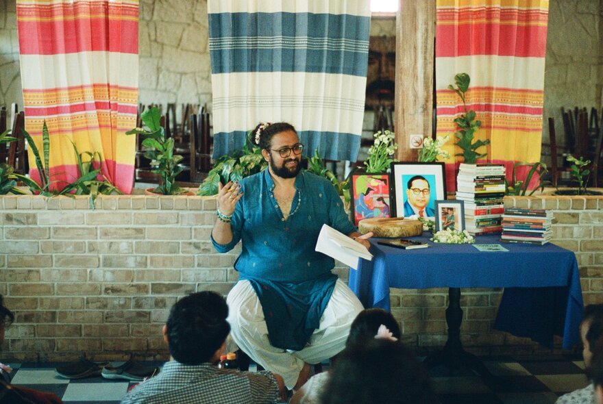A man sitting cross-legged in front of a small group of people with a table containing books beside him and coloured material hanging behind him.