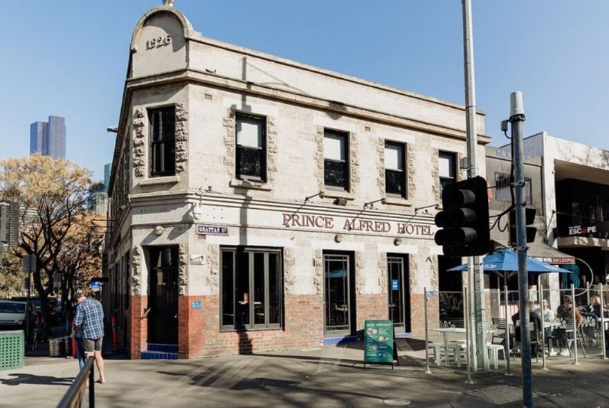 The building exterior of the Prince Alfred Hotel, on a street corner, daytime, with a person walking on the street in front of it.