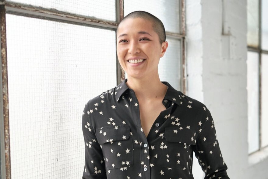 Jess Ho with a buzz cut near a large frosted window in a warehouse. 