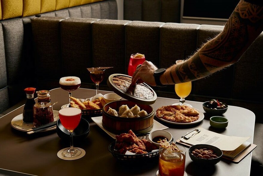 Someone taking the lid off a steamer full of dumplings at a table laden with lots of food, drinks and condiments. 