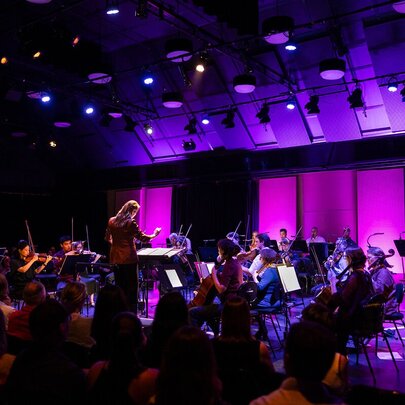Orchestra playing under purple lights, with conductor in front.