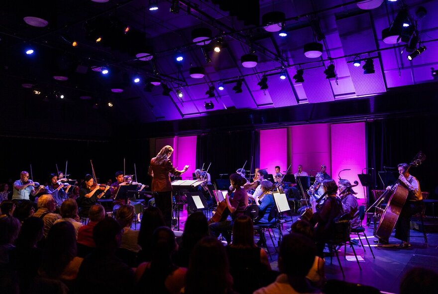 Orchestra playing under purple lights, with conductor in front.