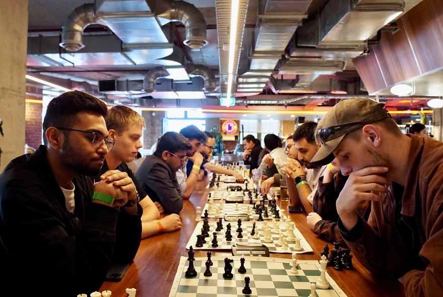 A social chess gathering where a long table of people are playing chess matches in an indoor setting.