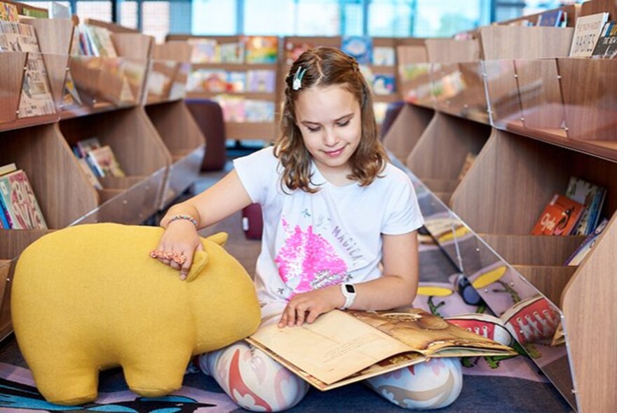 A young child sitting on the floor in the children's section of a library reading a picture book, her hand resting on a large stuffed yellow toy.