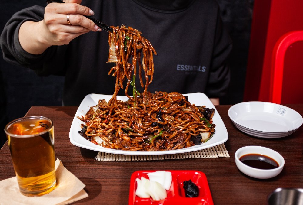 Person picking up seafood black bean sauce noodles with chopsticks, with a glass of beer, garnish and soy sauce on the side.