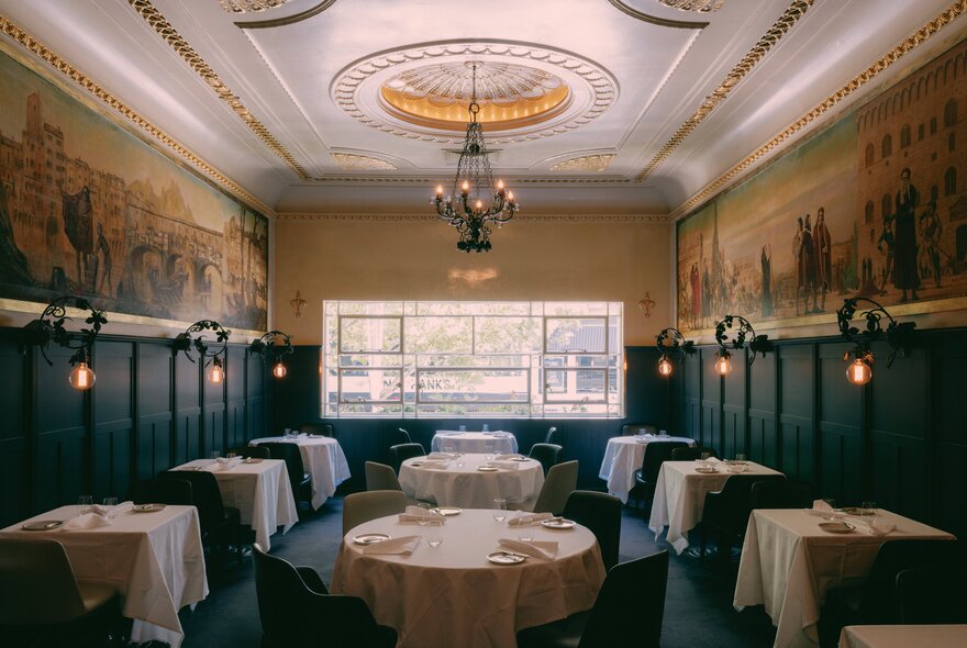 An elegant dining room with white-clothed tables, dark wood panelling, an ornate ceiling with a central chandelier, and large classical murals lining the upper walls.