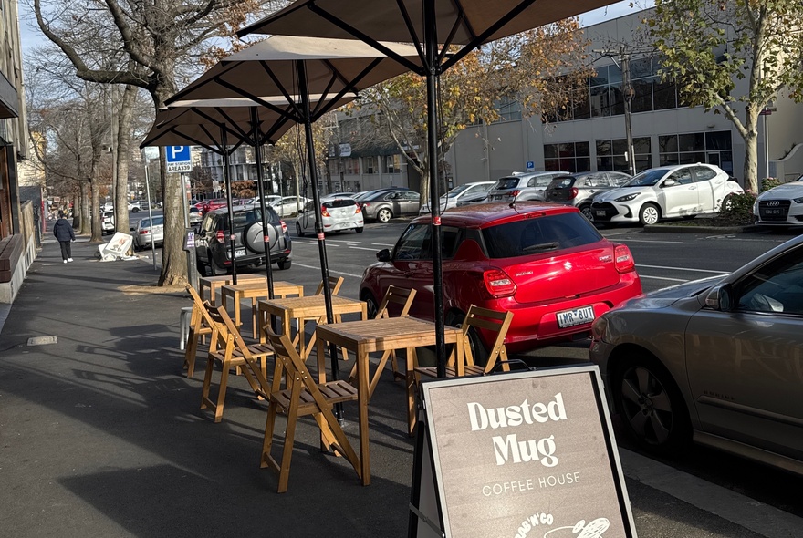 Cafe tables, chairs and umbrellas on a pavement with signage and parked cars.
