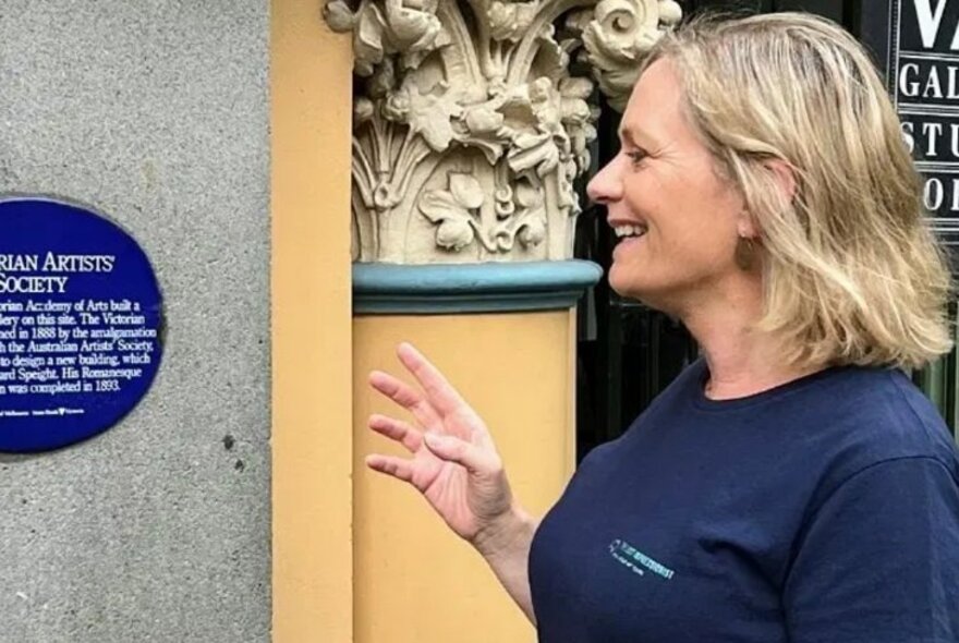 A smiling woman in a blue tour guide t-shirt, talking about a plaque on a concrete wall, alongside a column with elaborate carvings.