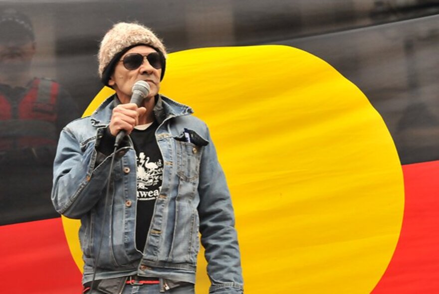Australian Aboriginal political activist Robbie Thorpe, holding a microphone in front of the Aboriginal flag.