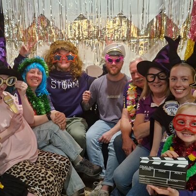 A group of people posing together in a large photo booth, wearing various costumes, accessories, and using props such as hats, masks, and glasses.