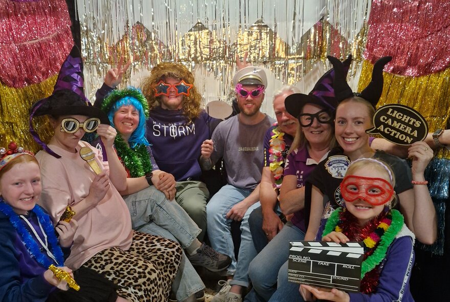 A group of people posing together in a large photo booth, wearing various costumes, accessories, and using props such as hats, masks, and glasses.