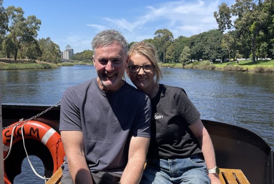 A smiling couple enjoying a sightseeing cruise in a boat along the Yarra River, with the river and trees in the background.