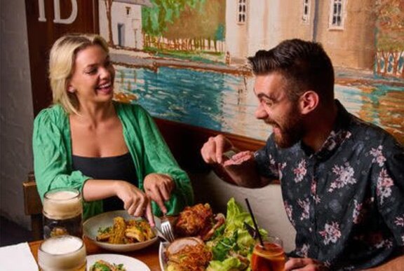 A couple eating at a restaurant with plates of food in front of them and a painted mural on the wall behind their table.