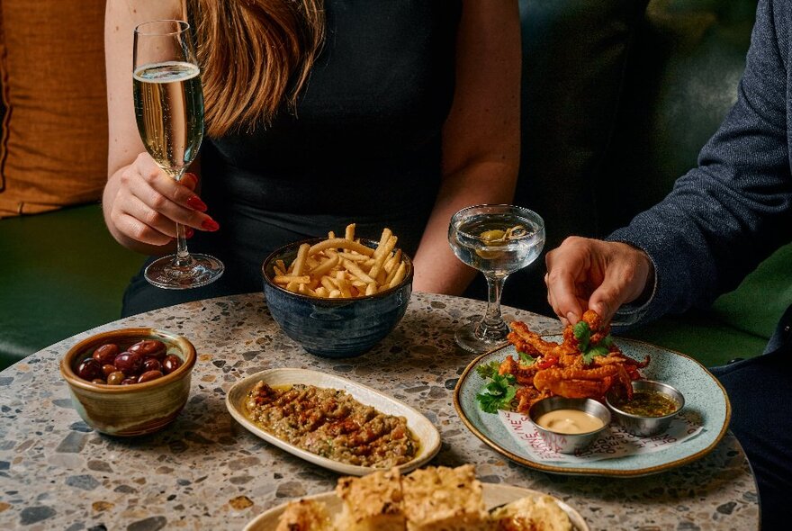 A small marble table filled with various share plates; two people seated around it with one holding a glass of wine. 