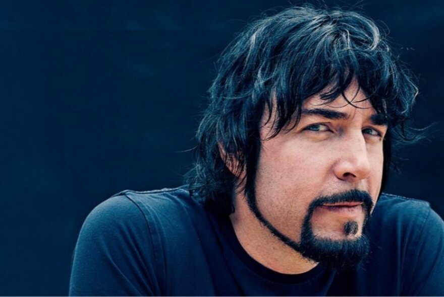 Bestselling dark fantasy author Jay Kristoff looking serious with tousled black hair, a black thin bead, black top against a black background.