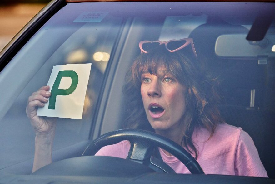 Movie still of a woman holding up a P probabionary licence sign while seated behind the wheel in a car.