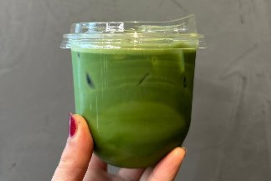 A matcha latte in a clear plastic cup and lid, held by a woman's hand with red nail polish. 