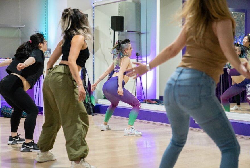 Dancers lined up in a row behind an instructor in front of a mirror, with knees bent and behinds sticking out.