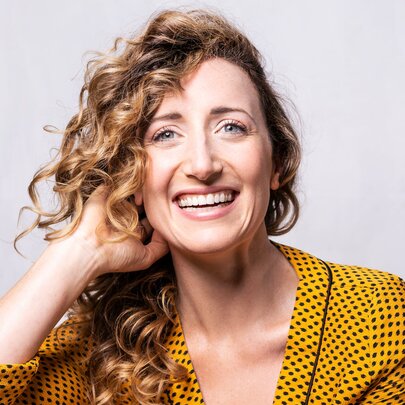 Australian actress and singer Elise McCann, smiling into the camera, with curly dark blonde hair, wearing a yellow jacket with small black polka dots. 