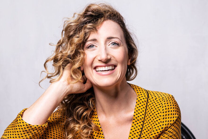 Australian actress and singer Elise McCann, smiling into the camera, with curly dark blonde hair, wearing a yellow jacket with small black polka dots. 