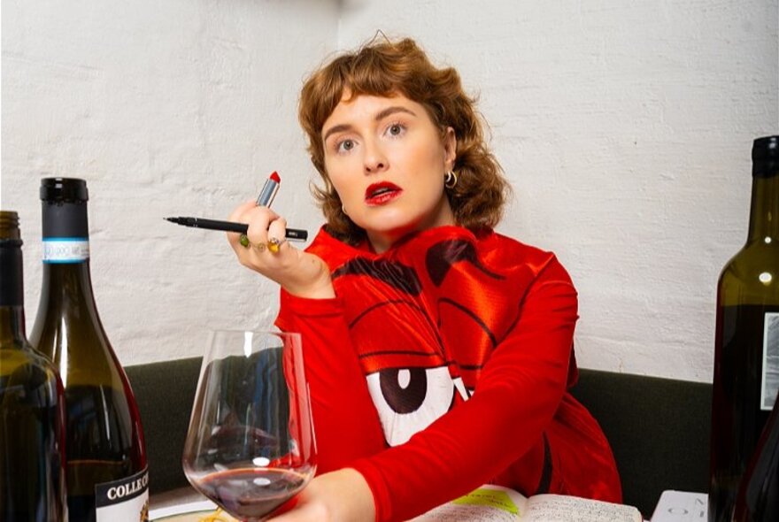 A woman wearing a red dress seated at a dining table with a glass of wine and bottle of wine around her, holding an open red lipstick in her hand and looking directly at the viewer.