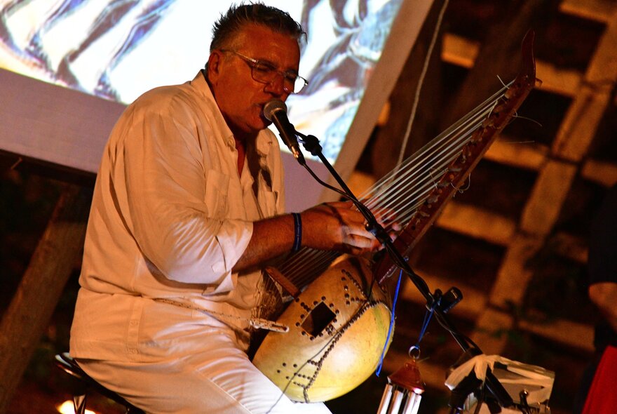 Italian musician, Fulvio Cama, wearing a white shirt and pants and holding a traditional Italian instrument, on stage with a microphone.