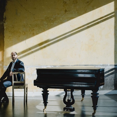 Musician Kirill Gerstein seated facing away from a grand piano in a room with sunlight beaming through windows. 