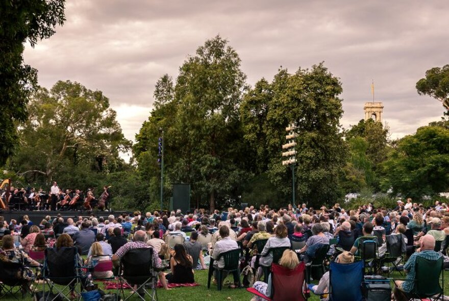 People seated in low chairs or on the lawns of the Royal Botanic Gardens watching an outdoor twilight performance. 