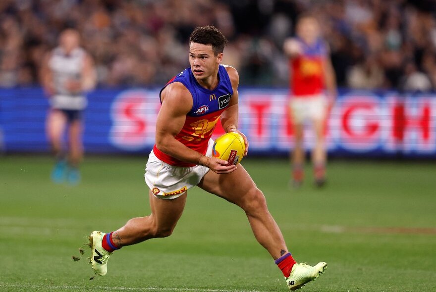 A Brisbane Lions player runs and turns with a football on a football field, out of focus players on the background. 