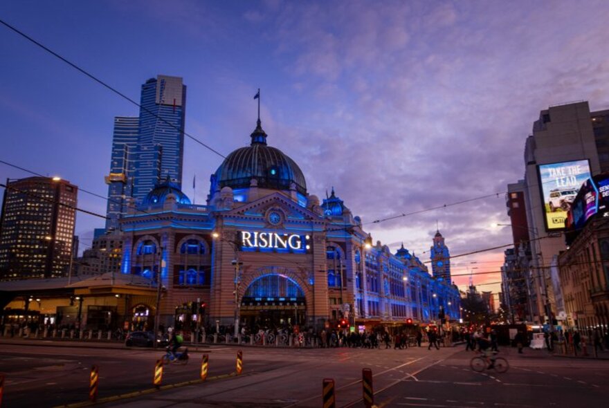 Flinders Street Station lit up in blue at dusk during Melbourne's RISING festival, with city buildings in the background. 