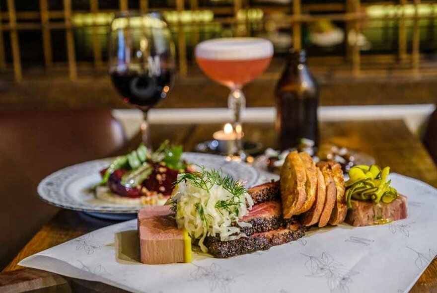 Charred meat, crudites and other cheese board items on a plate in front of a cocktail