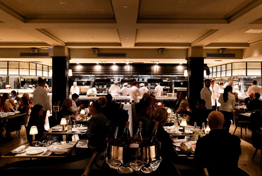 A busy restaurant with dim lighting and an open kitchen with chefs cooking in the background.