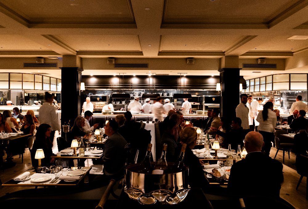 A busy restaurant with dim lighting and an open kitchen with chefs cooking in the background.
