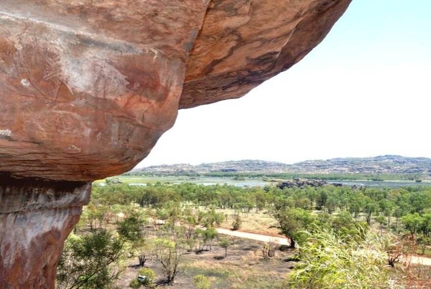 A large, reddish rock overhang with Aboriginal rock art visible on it, stands out in the foreground, with trees and hills under a clear sky in the background.