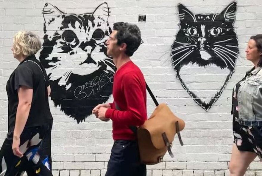People walking past stencil artwork of cats on a white wall in a Melbourne laneway.