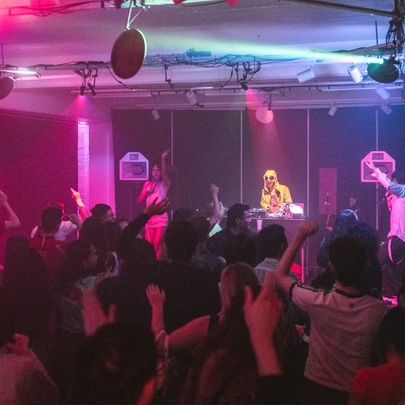 A crowded indoor nightclub scene with a DJ performing on stage under purple and blue lighting and people dancing.